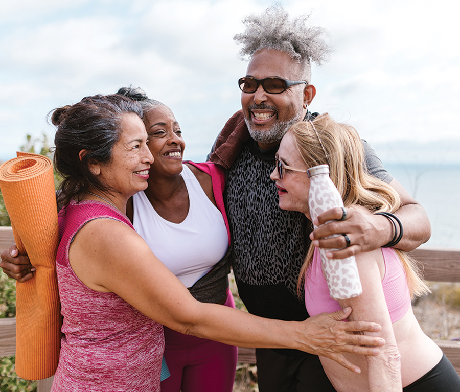 A group of four people, dressed in activewear, smiling and embracing each other outdoors. One person holds an orange yoga mat, and another holds a white water bottle. The background shows a scenic outdoor setting with a wooden fence and distant water view.