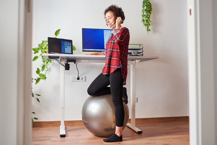 Person incorporating movement into the workday by using a stability ball and adjustable standing desk in a home office.