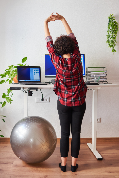 Person stretching while using a sit-stand desk in a home office workspace, demonstrating the importance of regular position changes throughout the workday.